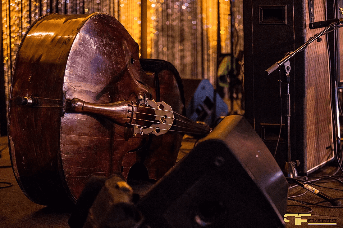 Upright bass resting by a floor wedge monitor and mic stand, sequin curtain behind