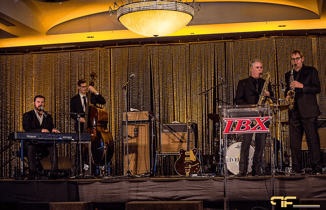 Full band on skirted stage: Roland keys, upright bass, saxes, IBX acrylic podium, gold curtain