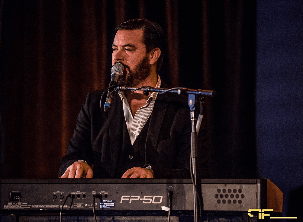 Bearded keyboardist singing at a Roland FP-50 stage piano, dark drape behind