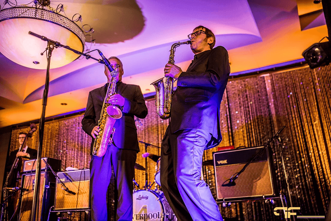 Saxophonists on the band stage under a crystal chandelier, Vox amp and drum kit behind