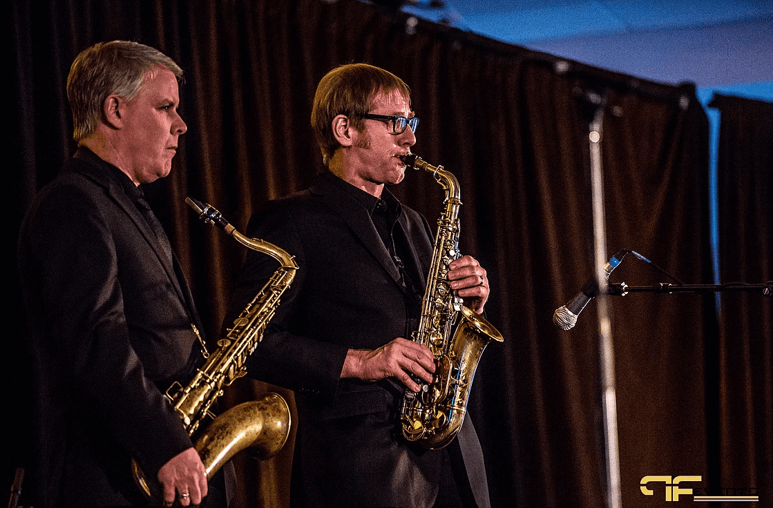 Two saxophonists performing in front of dark brown stage drape with a vocal mic stand