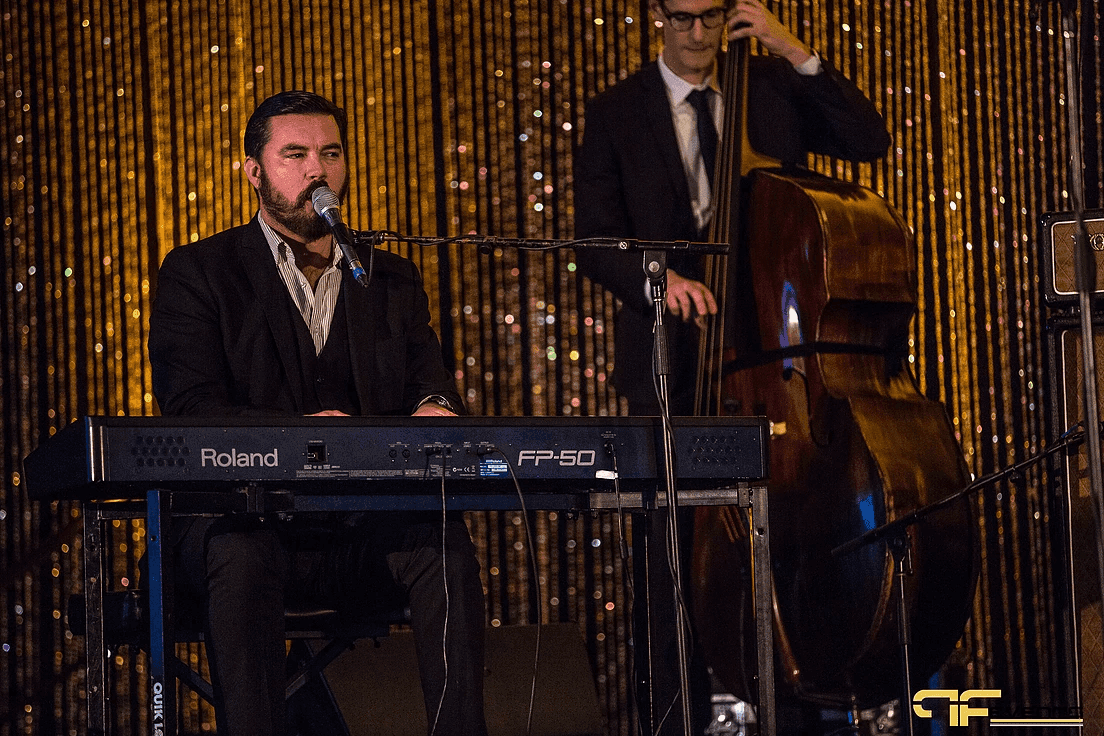 Keyboardist singing at a Roland FP-50 with upright bassist behind, gold sequin backdrop