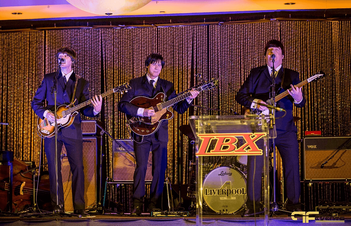 Four-piece Beatles tribute band on stage behind a clear acrylic podium with the IBX logo