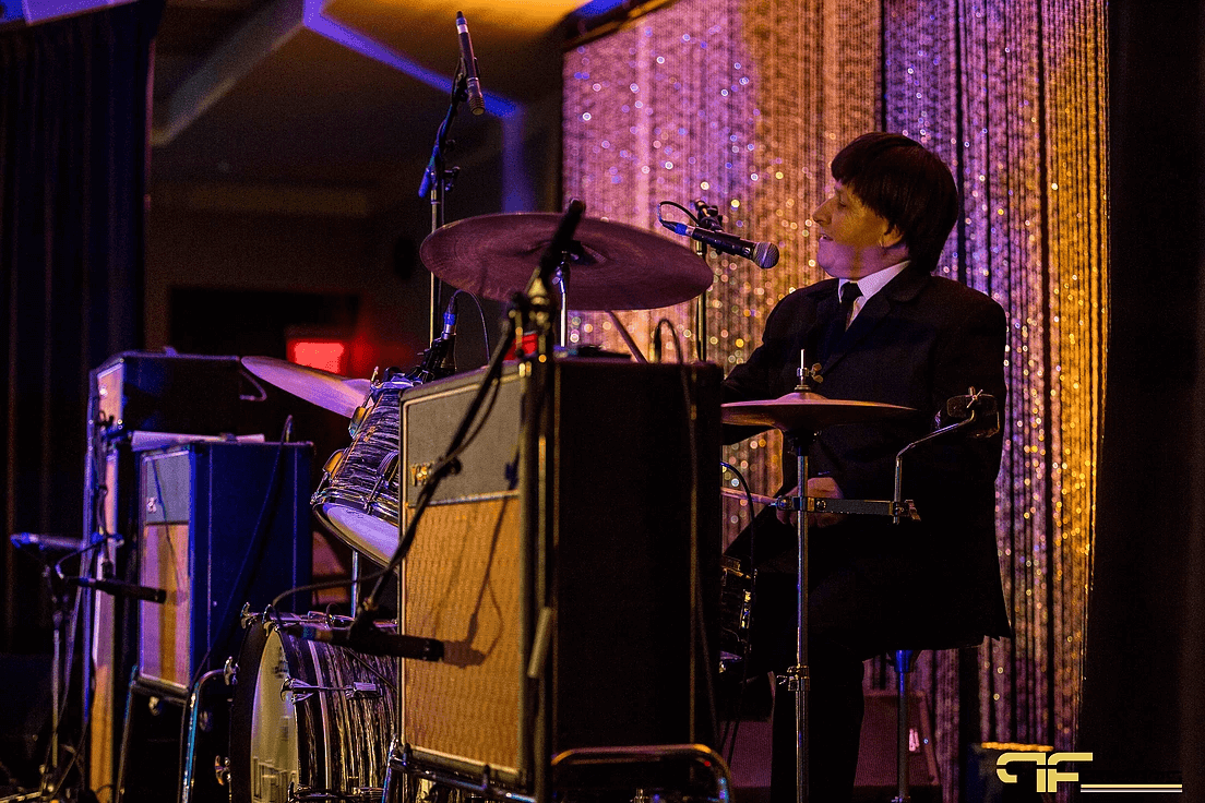 Tribute band drummer behind Vox amps and vocal mics against a sparkling strand curtain
