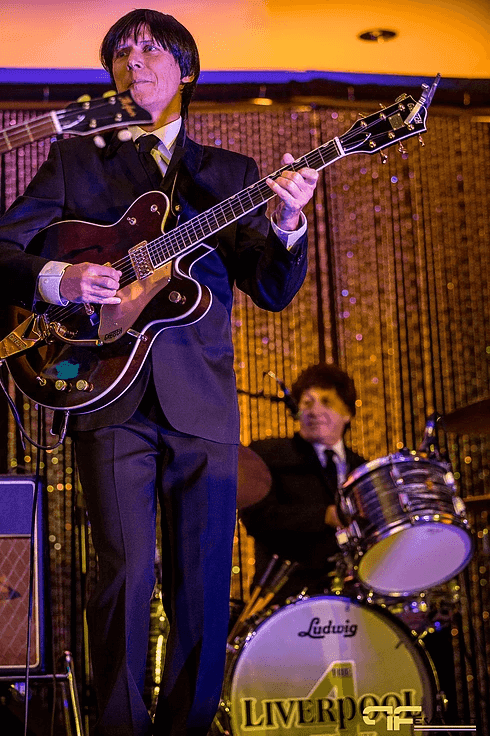 Beatles-tribute guitarist in dark suit on stage, gold sequin curtain and Ludwig kit behind