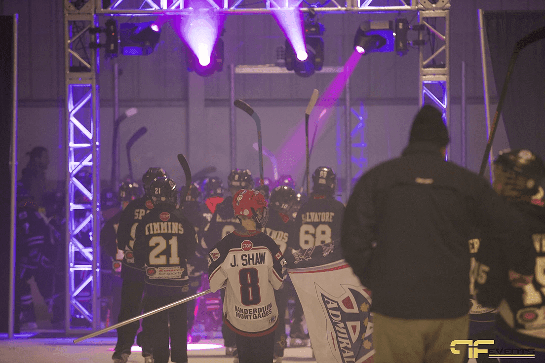 Players with sticks raised file out under the truss arch as magenta beams fire down
