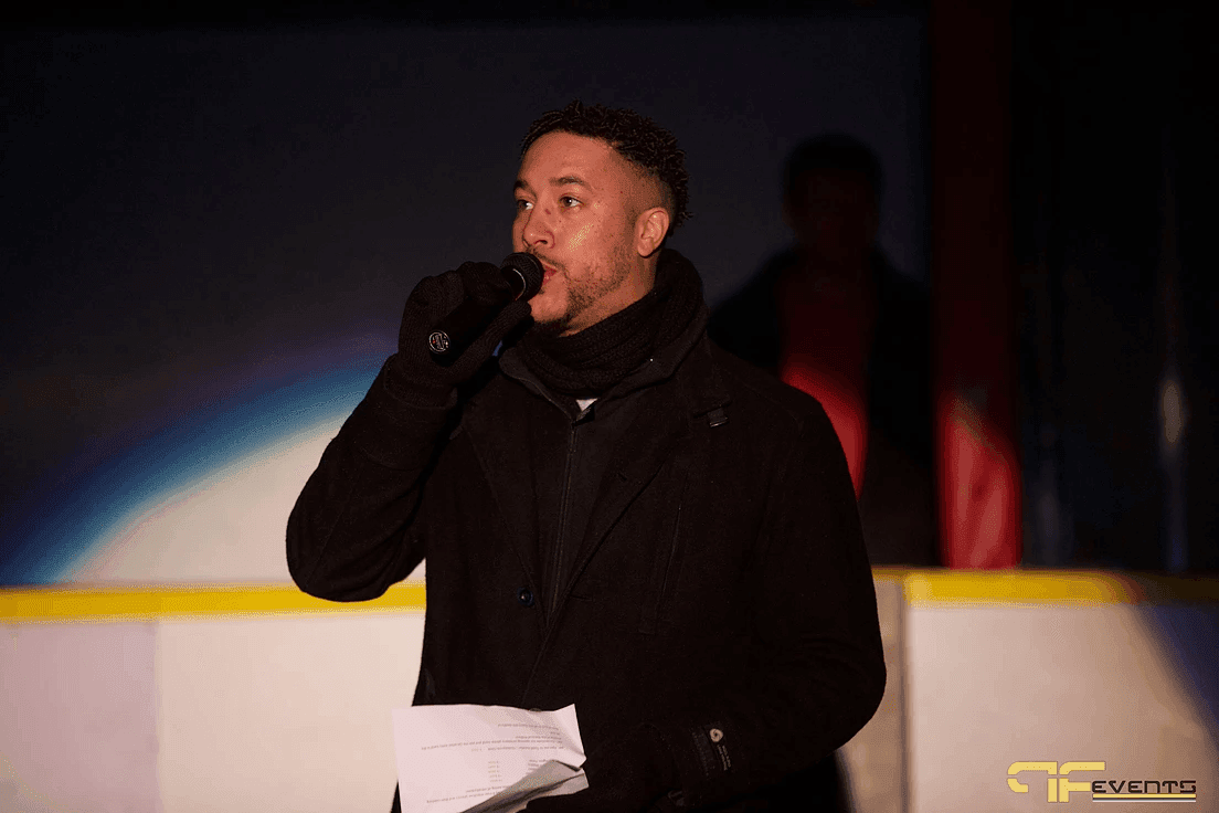MC in a black coat announcing into a handheld mic at the rink boards