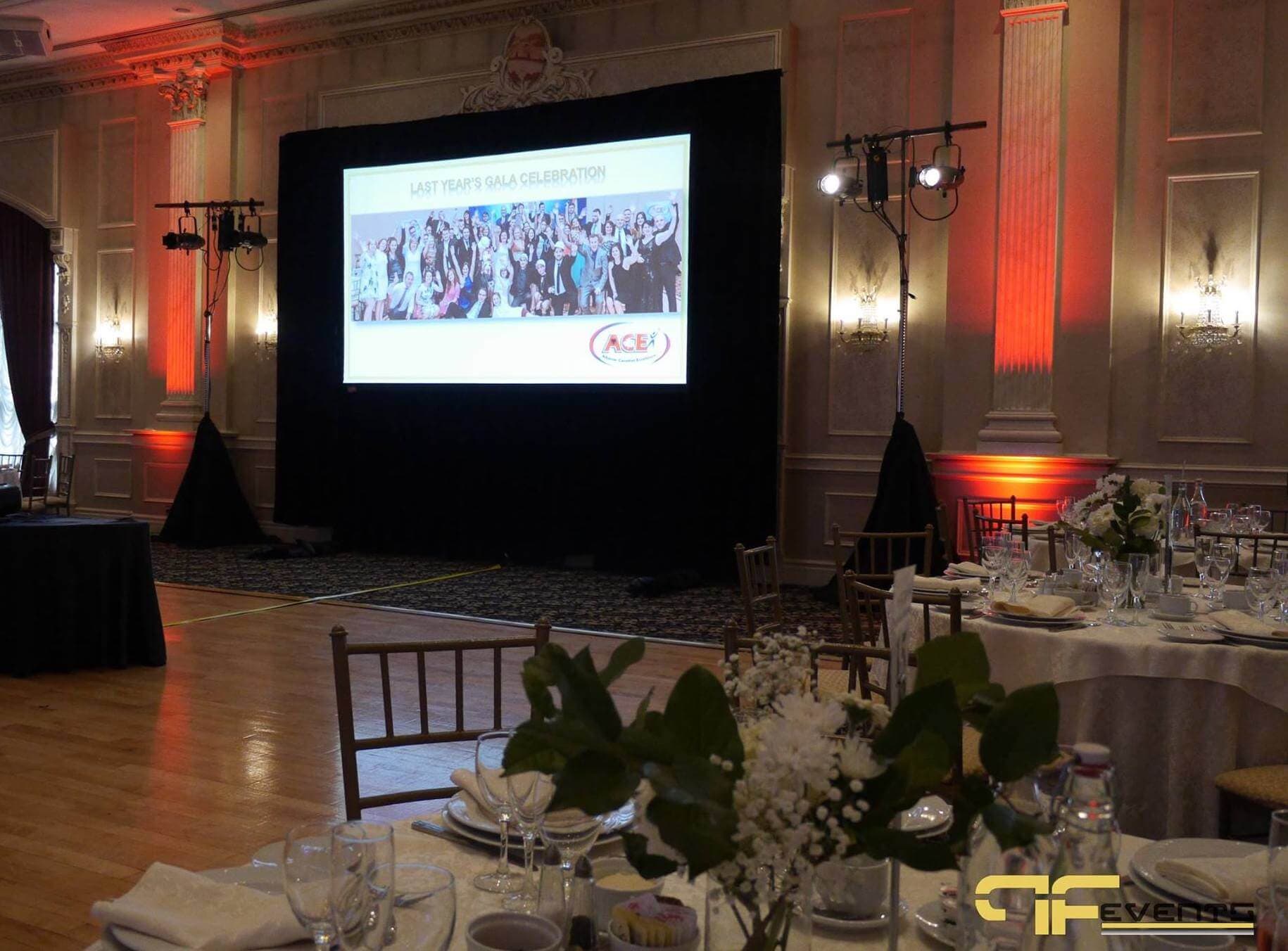 Projection screen in black drape flanked by spotlight stands and red wall uplighting