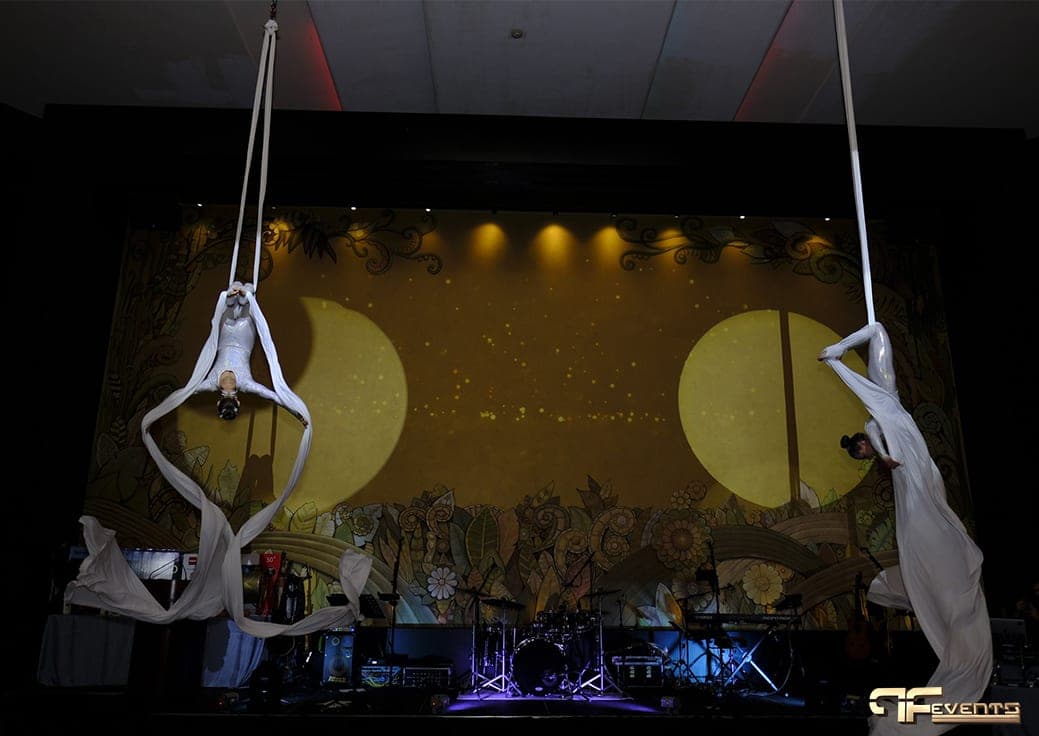 Two aerialists on white silks framing yellow moon projections on the stage
