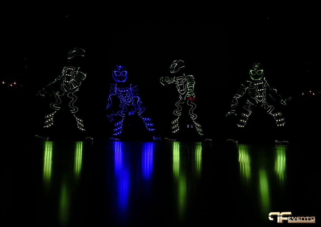 Four LED-suit dancers glowing blue and green, reflected in the dance floor