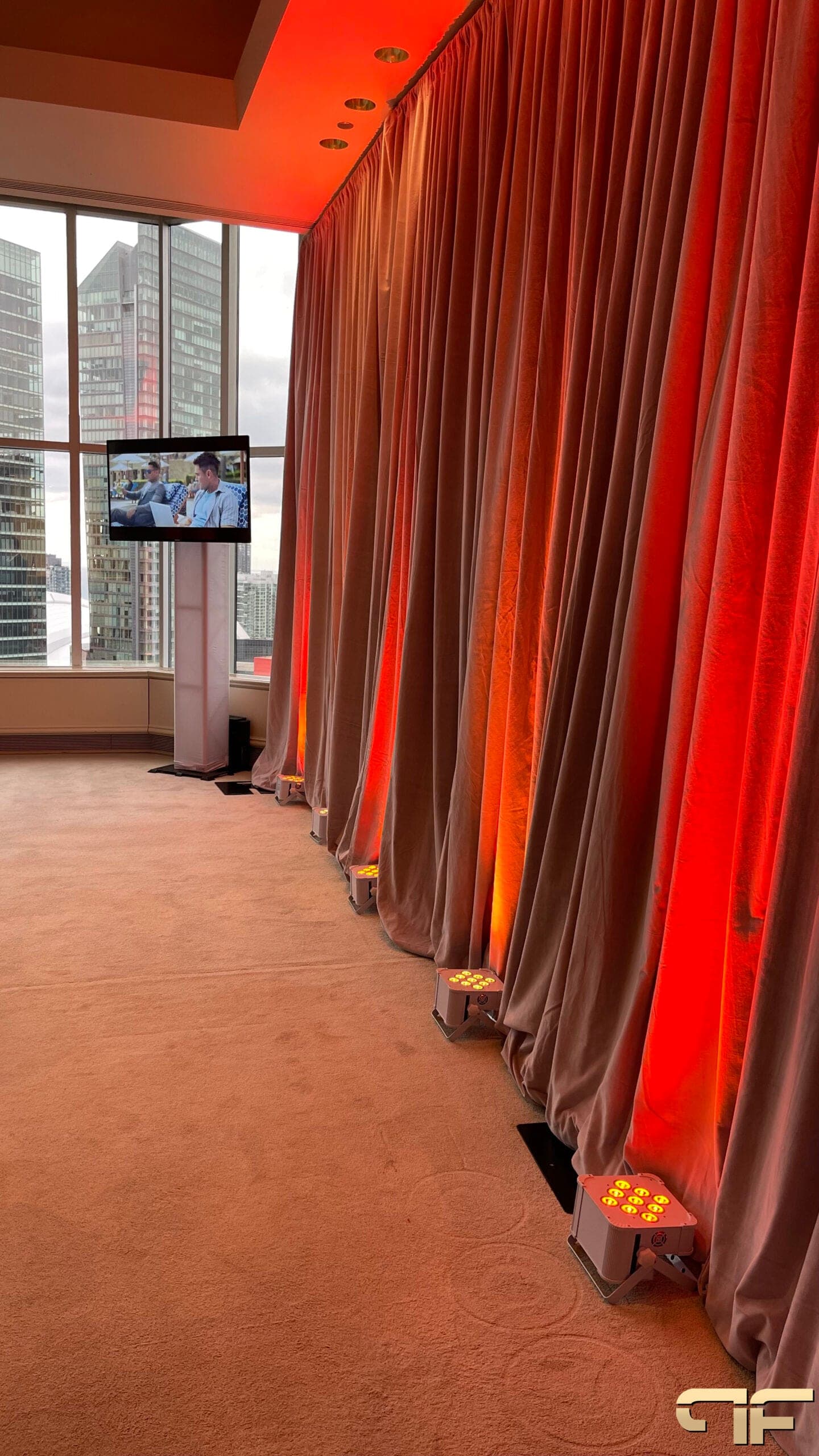 Velour drape wall washed orange by a row of floor uplights, TV totem by the window