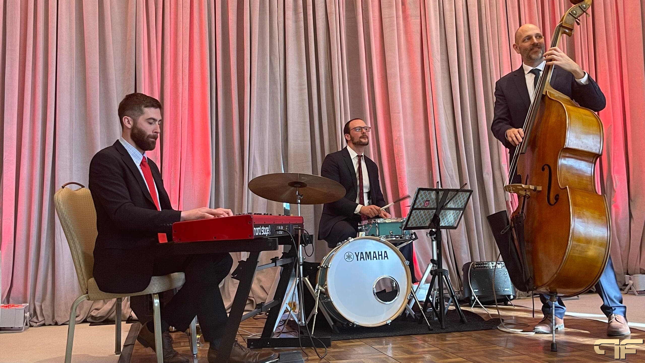 Jazz trio on keys, Yamaha drums and upright bass in front of pink-lit drapes