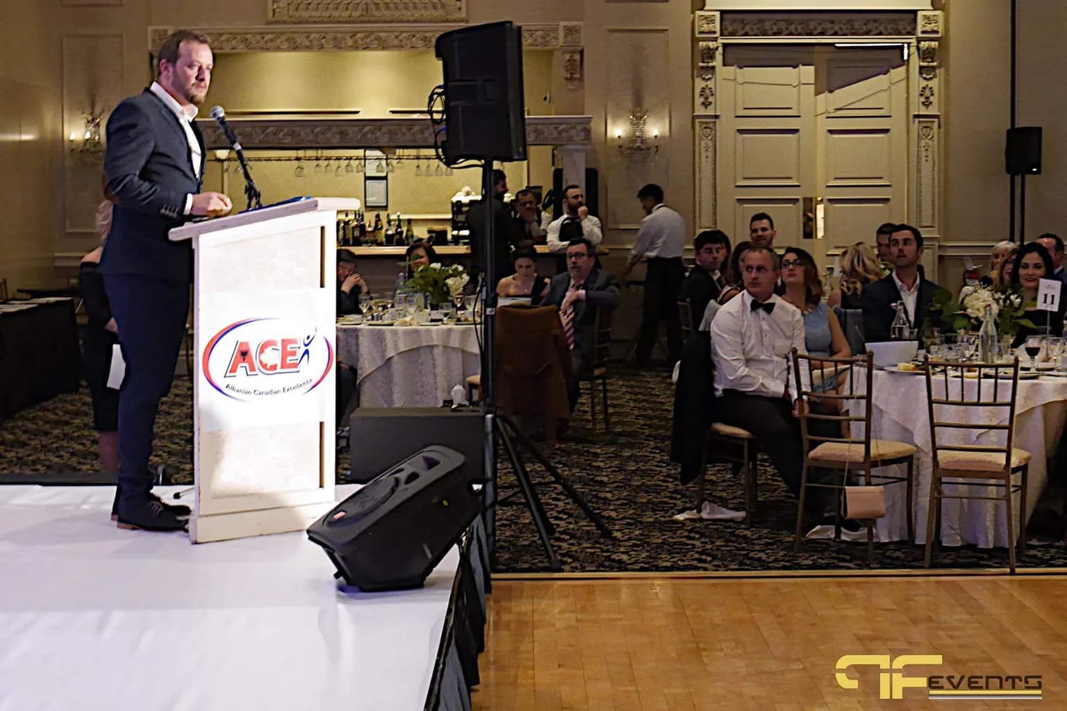 Presenter speaking at a branded lectern with stage speakers facing an audience at round tables