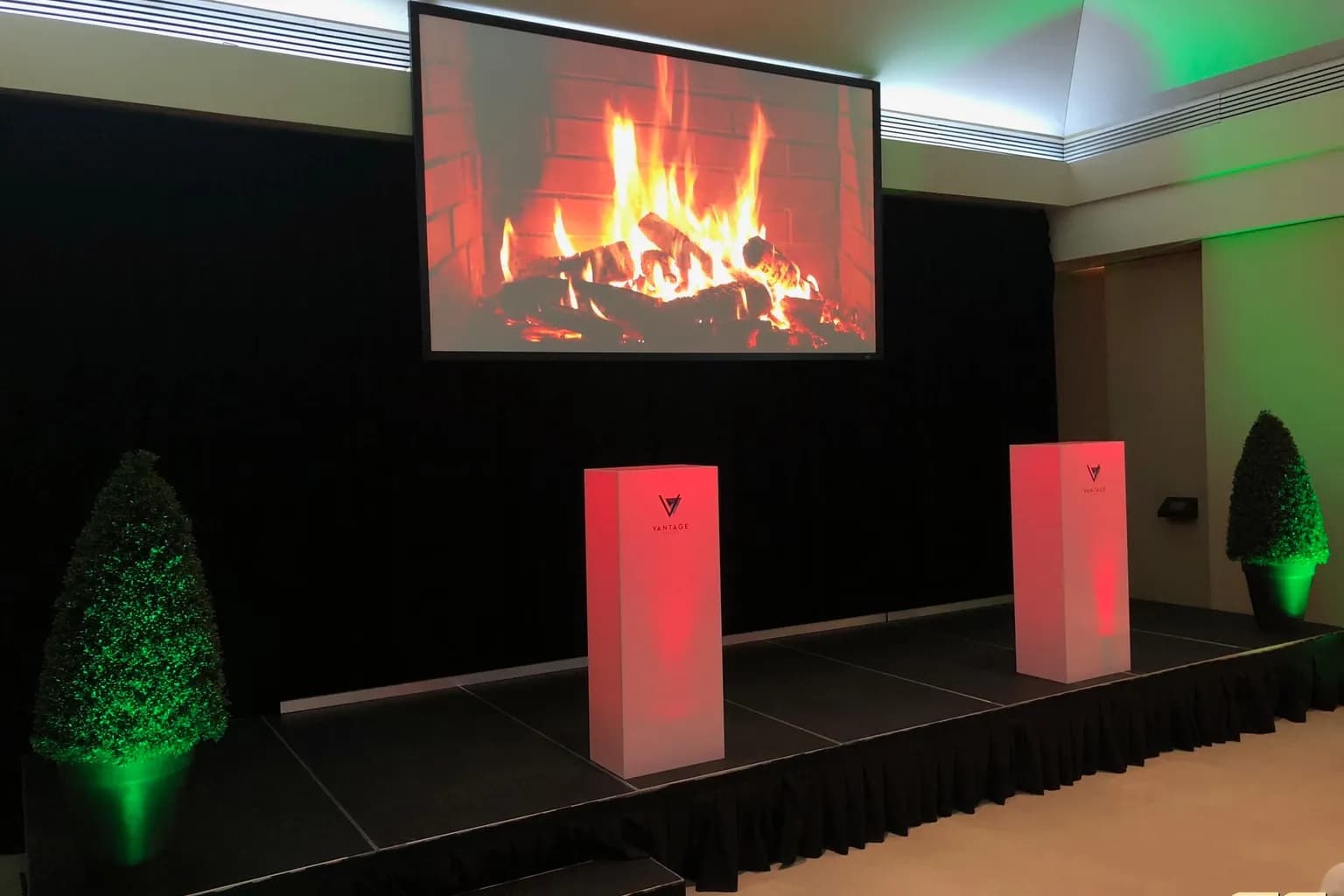 Stage with a fireplace video on the screen, lit trees, and green uplighting for a corporate holiday party