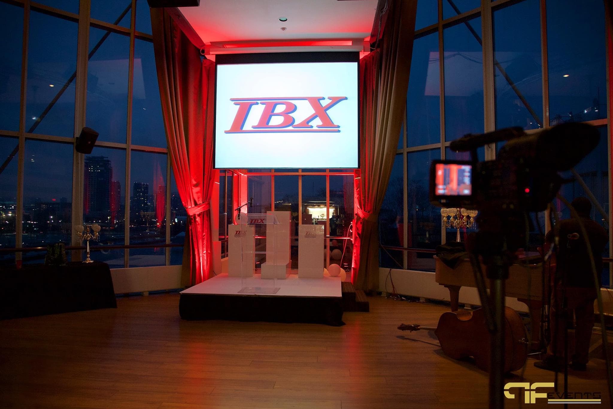 Projection screen with IBX logo over white stage riser, red-lit drapes, camera in foreground