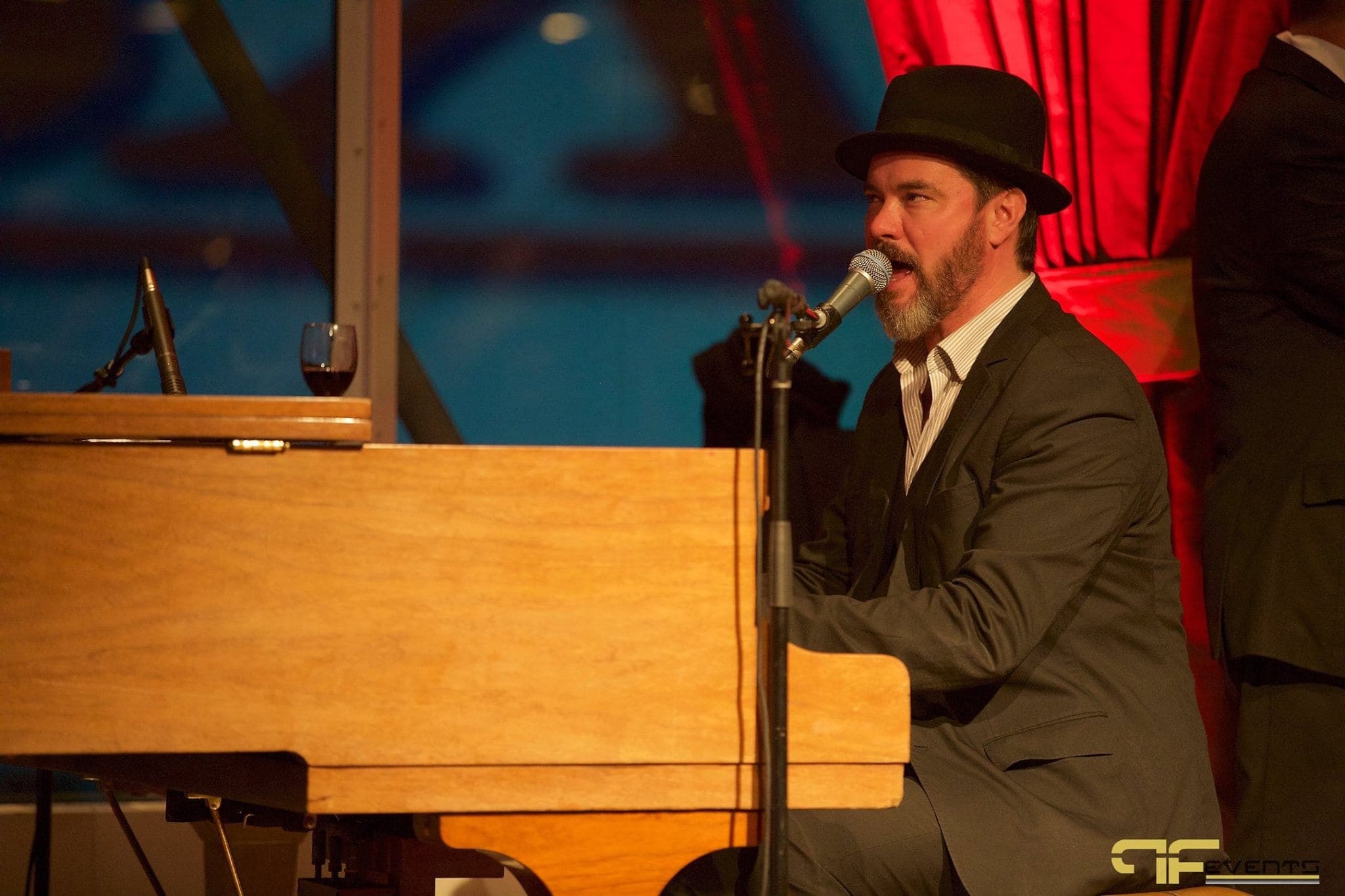 Pianist in a bowler hat singing at a baby grand piano beside red drapes