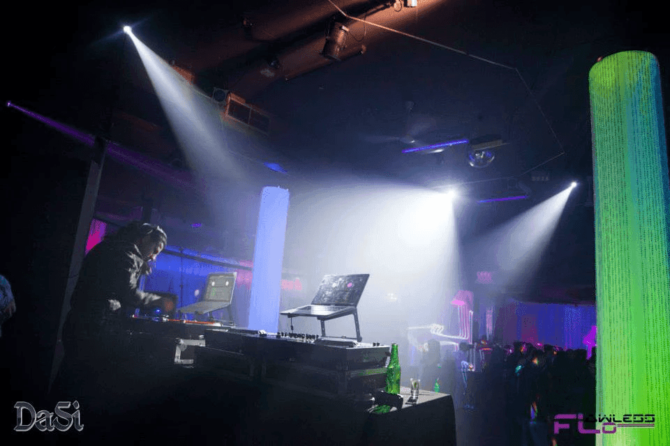 DJ booth under white moving-head beams in haze beside a glowing green column