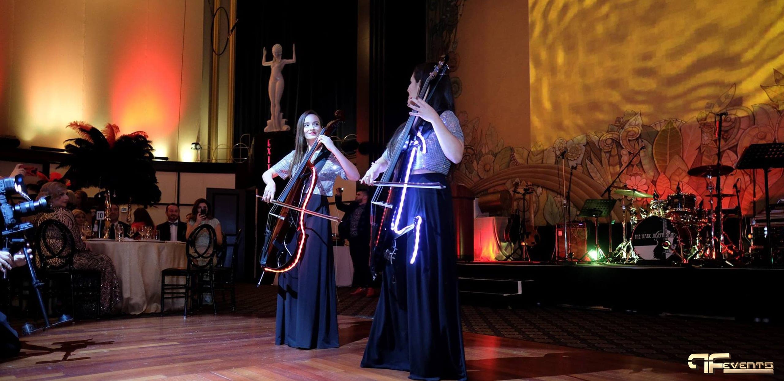 Two cellists with LED-outlined instruments under a gold rippling stage wash