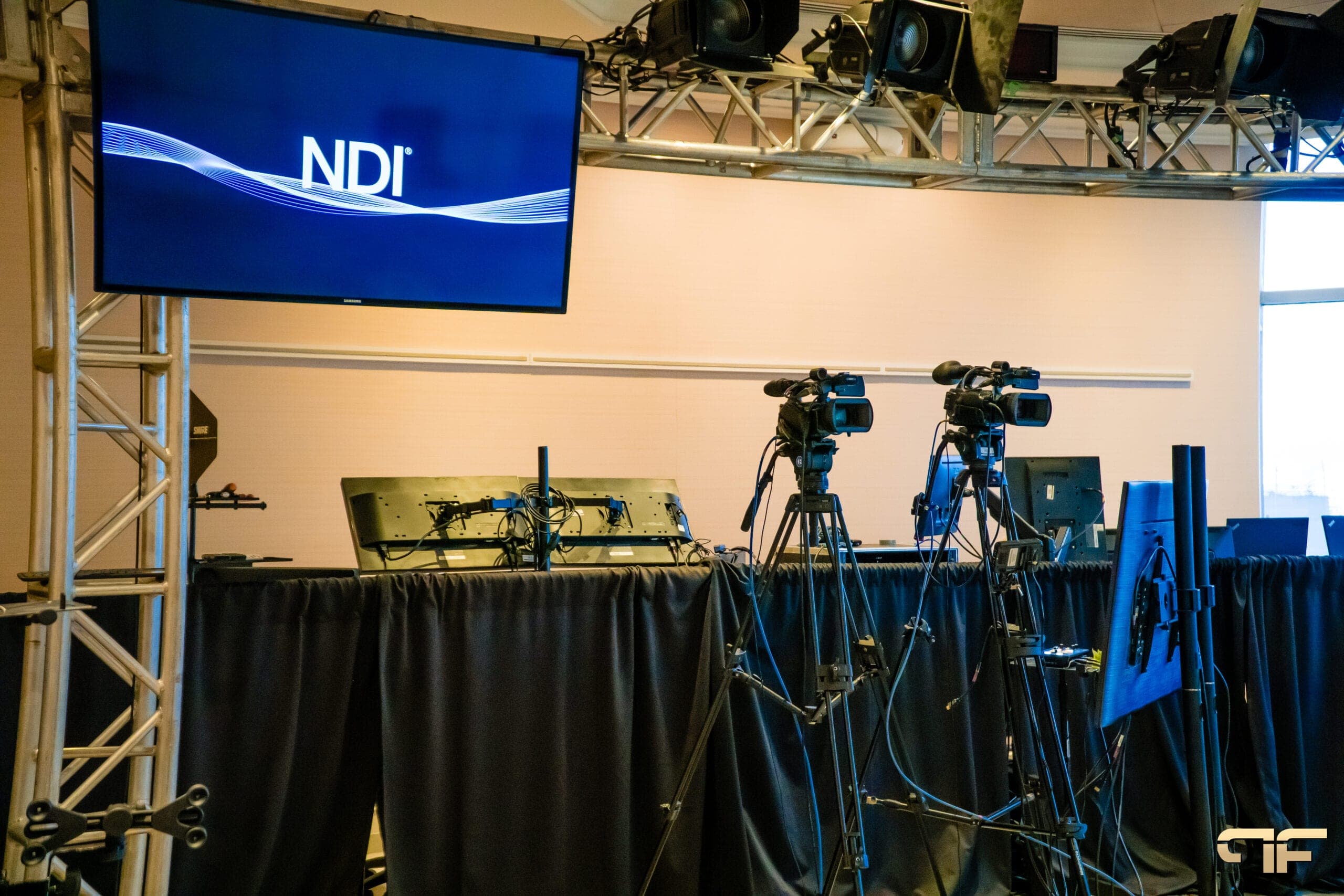Two video cameras on tripods beneath a truss grid and NDI screen