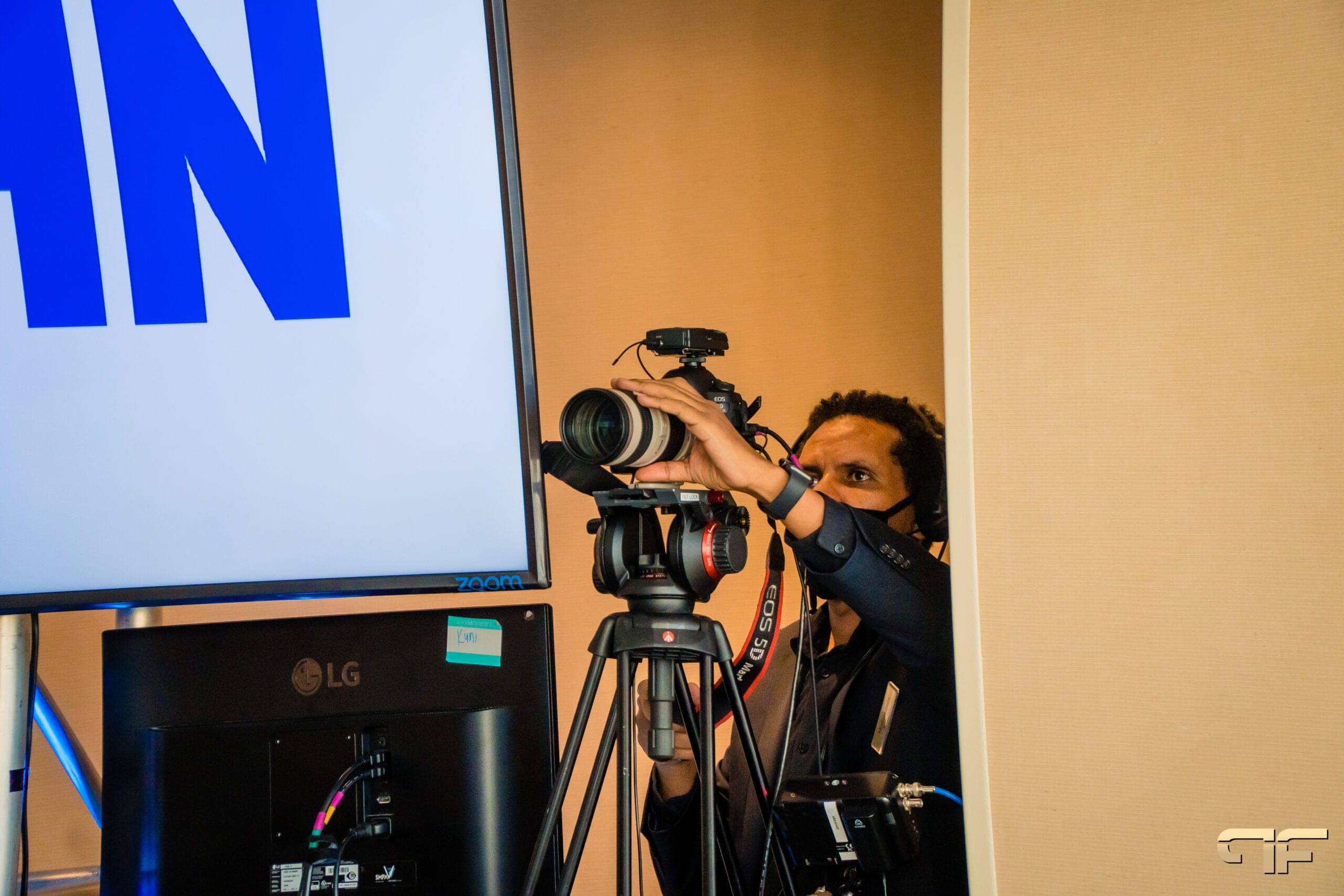 Camera operator adjusting a DSLR on a tripod beside a UHN-branded screen