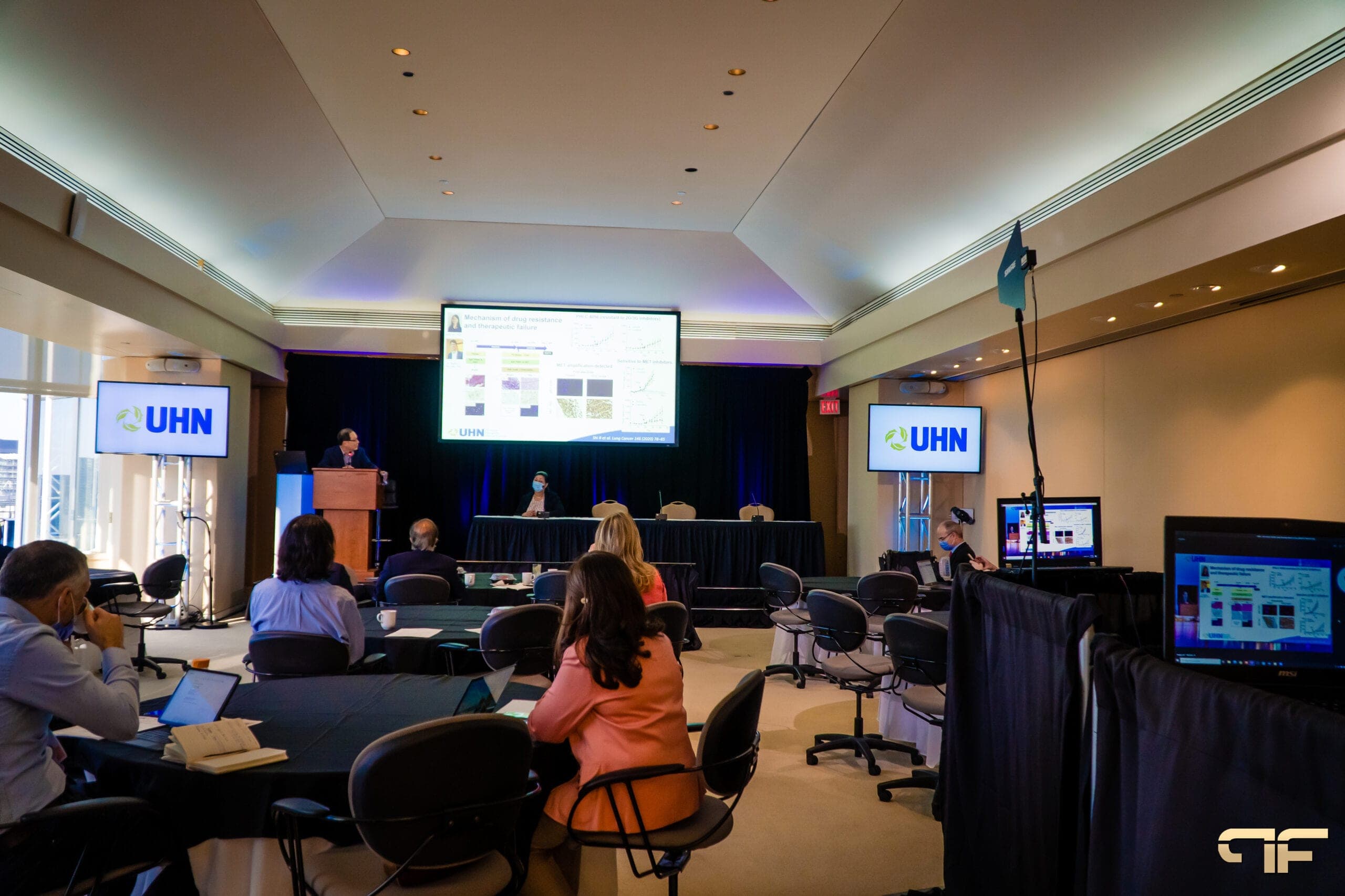 Distanced audience at round tables facing a presenter, UHN screens on truss