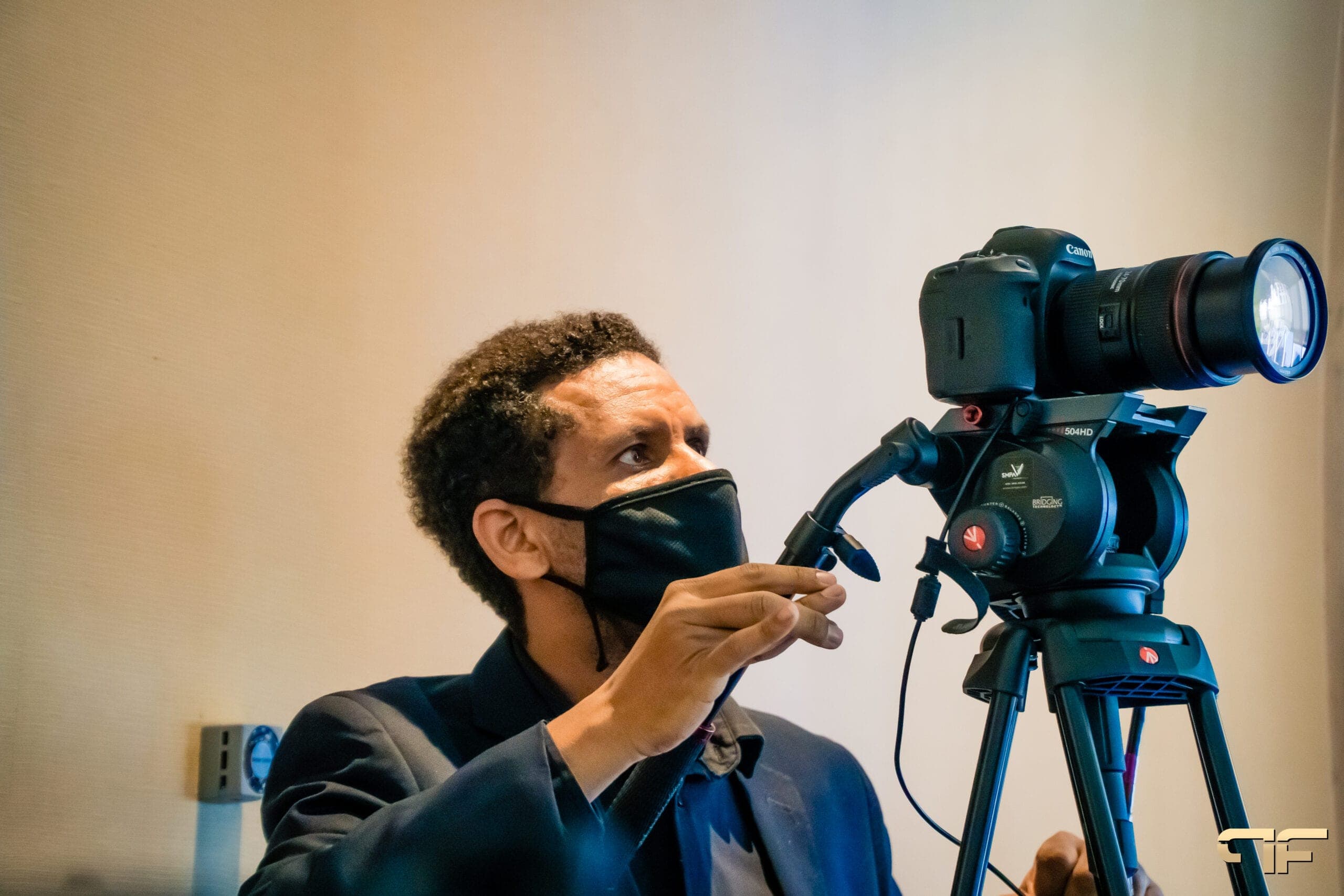 Camera operator in a mask adjusting a Canon camera on a fluid-head tripod